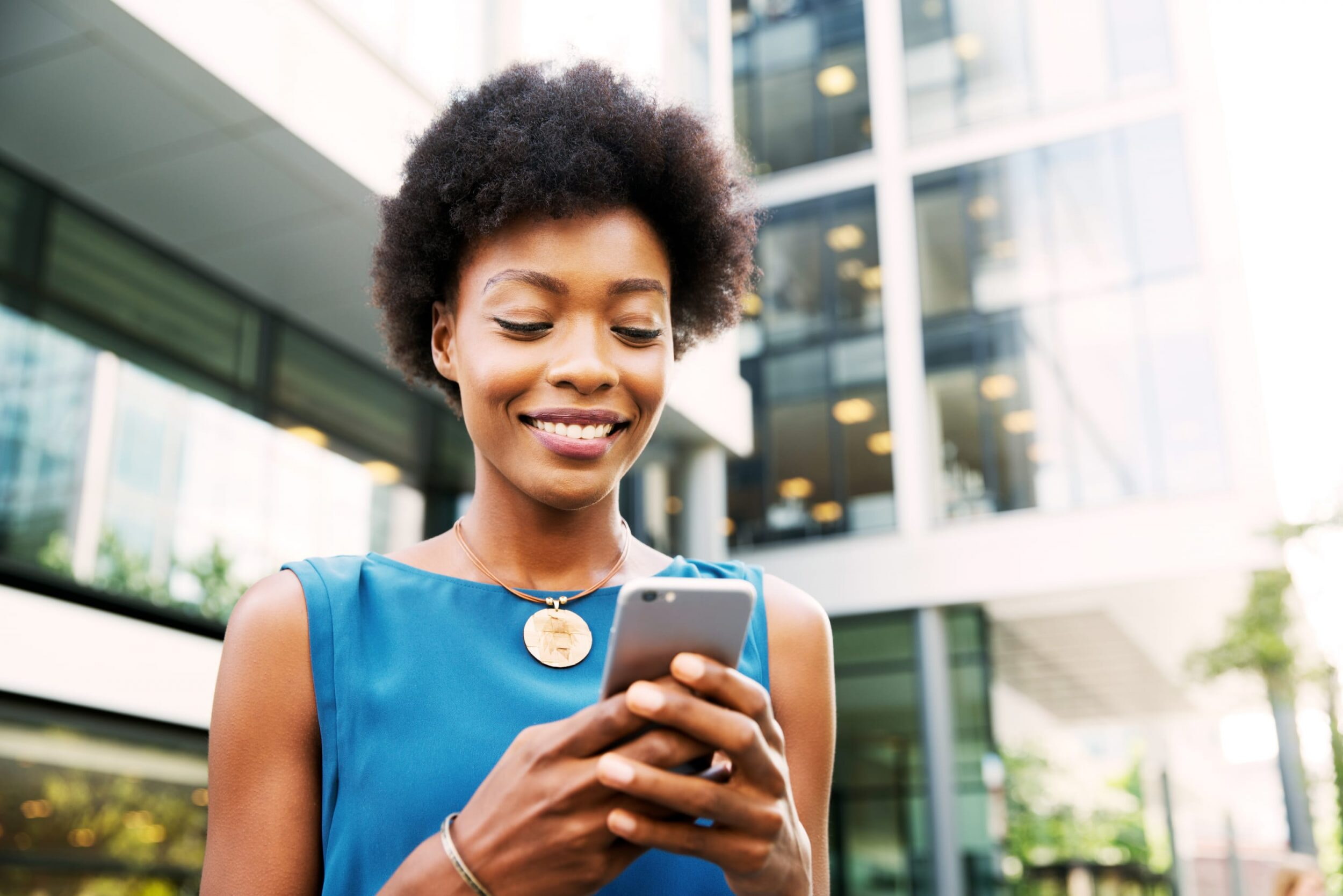 Eine Frau mit schwarzen, kurzen Haaren steht vor einem Gebäude. Sie ist an ihrem Smartphone und schreibt etwas.