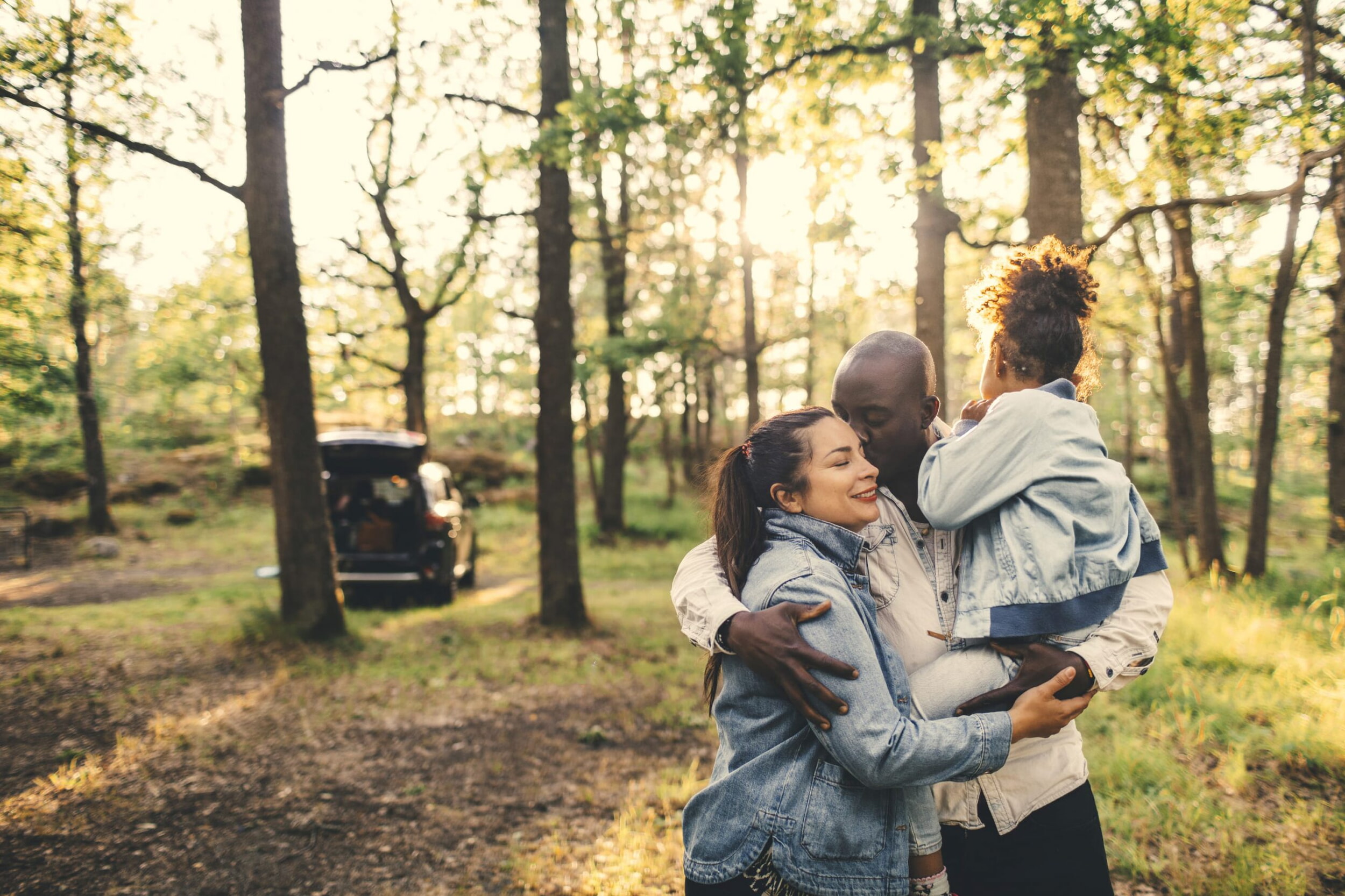 Una famiglia si trova nel bosco. L’uomo cinge la donna e tiene in braccio una bambina. C’è il sole.