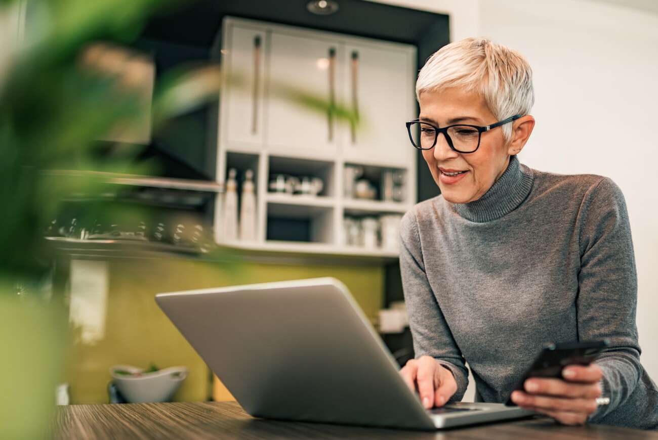 Eine Frau mit Haaren hält ein Smartphone in der Hand und schaut in den Laptop vor ihr.