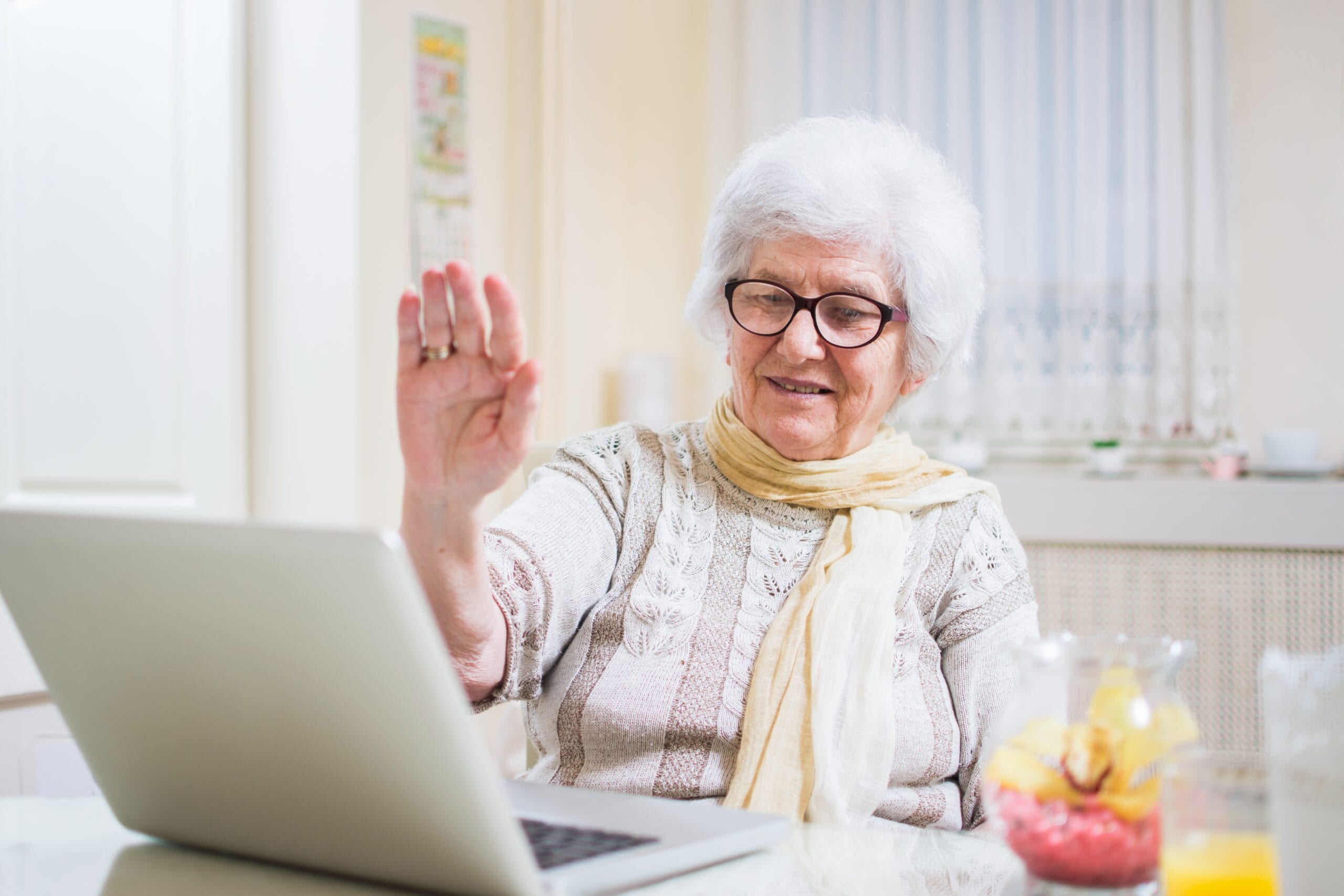 Gray-haired senior woman waving hand in front of laptop while having video call with her family members.
